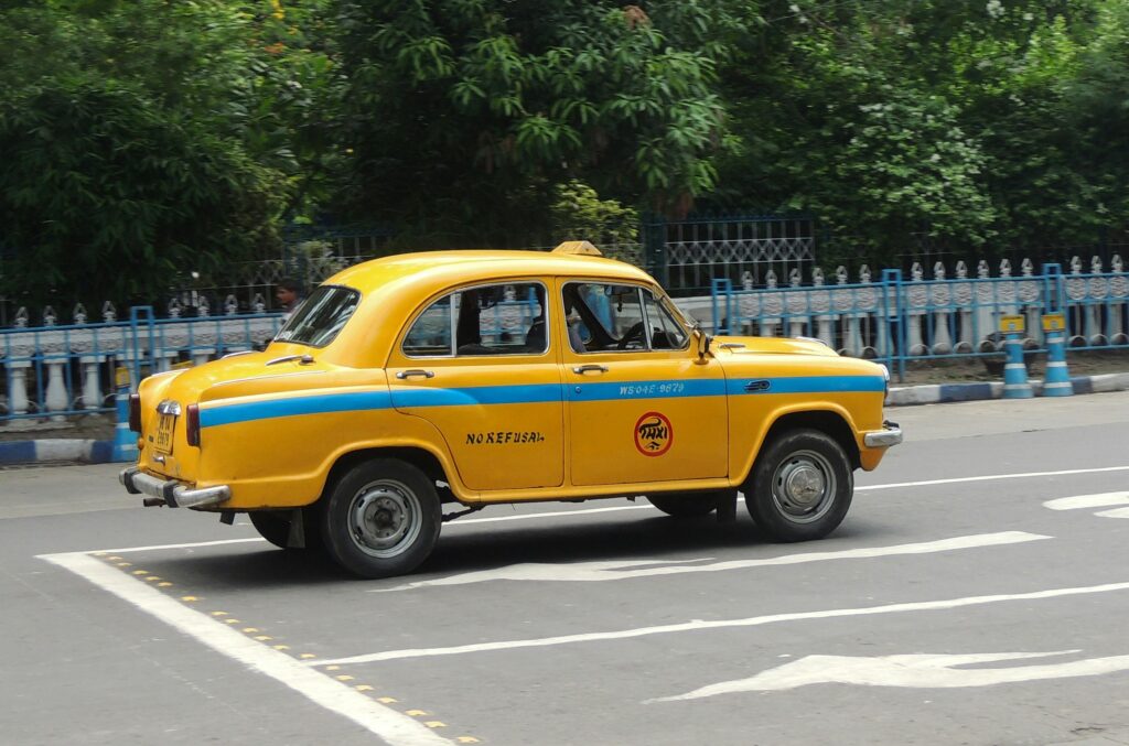 pexels-photo-26953406-26953406 Yellow taxi driving on the streets of Kolkata, India, showcasing urban life.